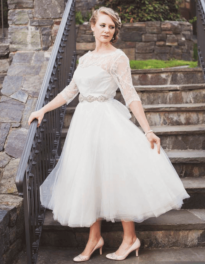 Bride in a tea-length lace wedding dress posing on stone steps with garden background.