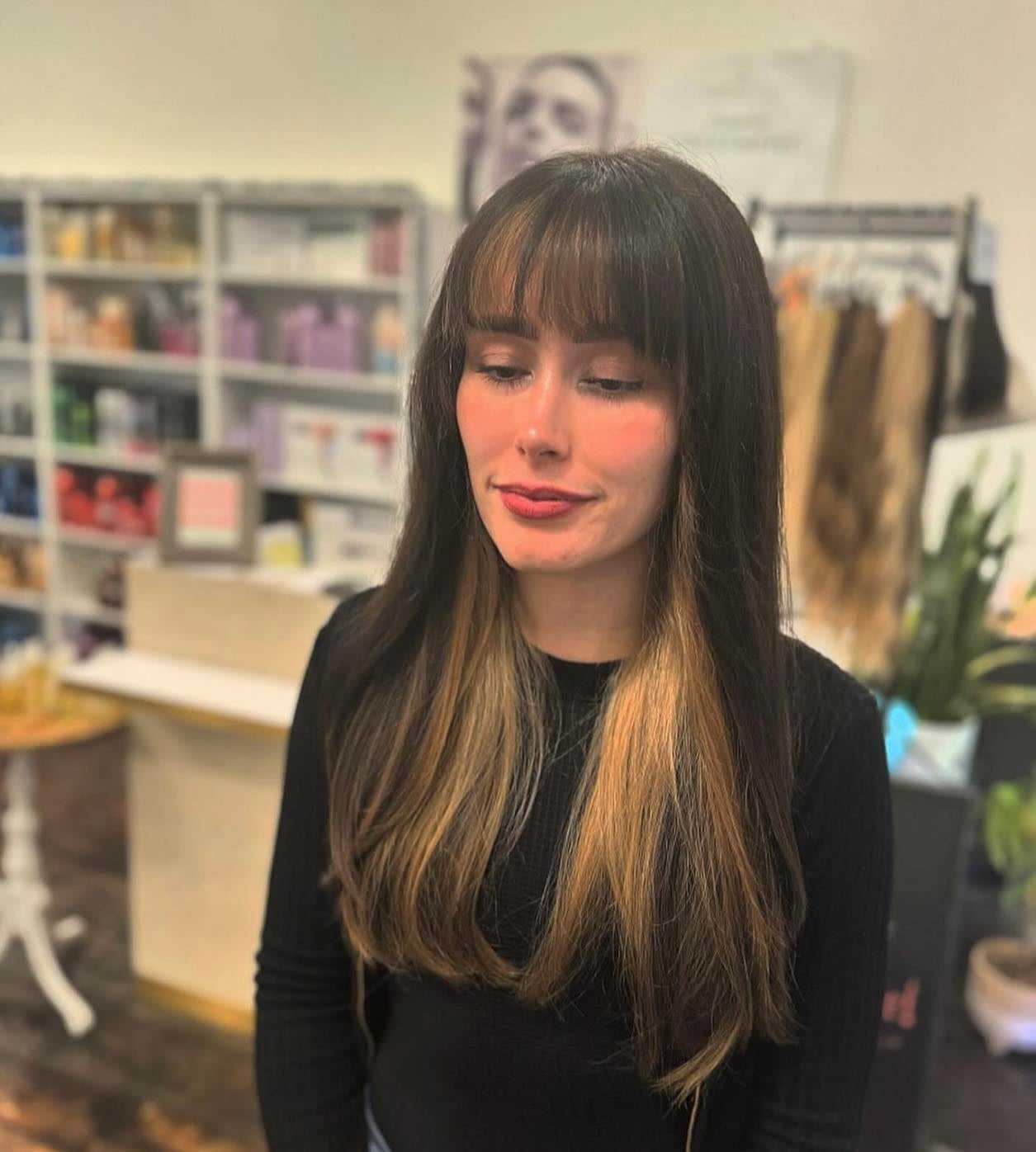 Woman with long, straight hair and bangs standing in a salon with products in the background.