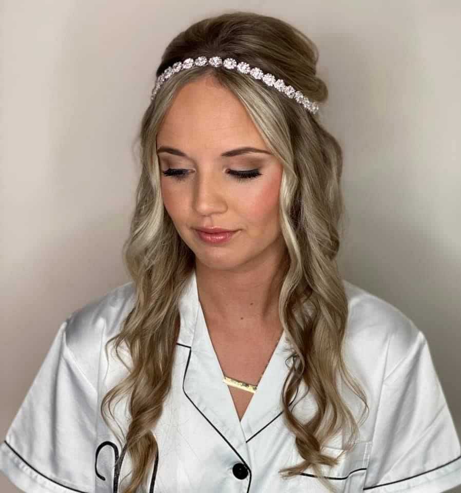 Young woman with curled hair, floral headband, and white blouse looking down against plain background.